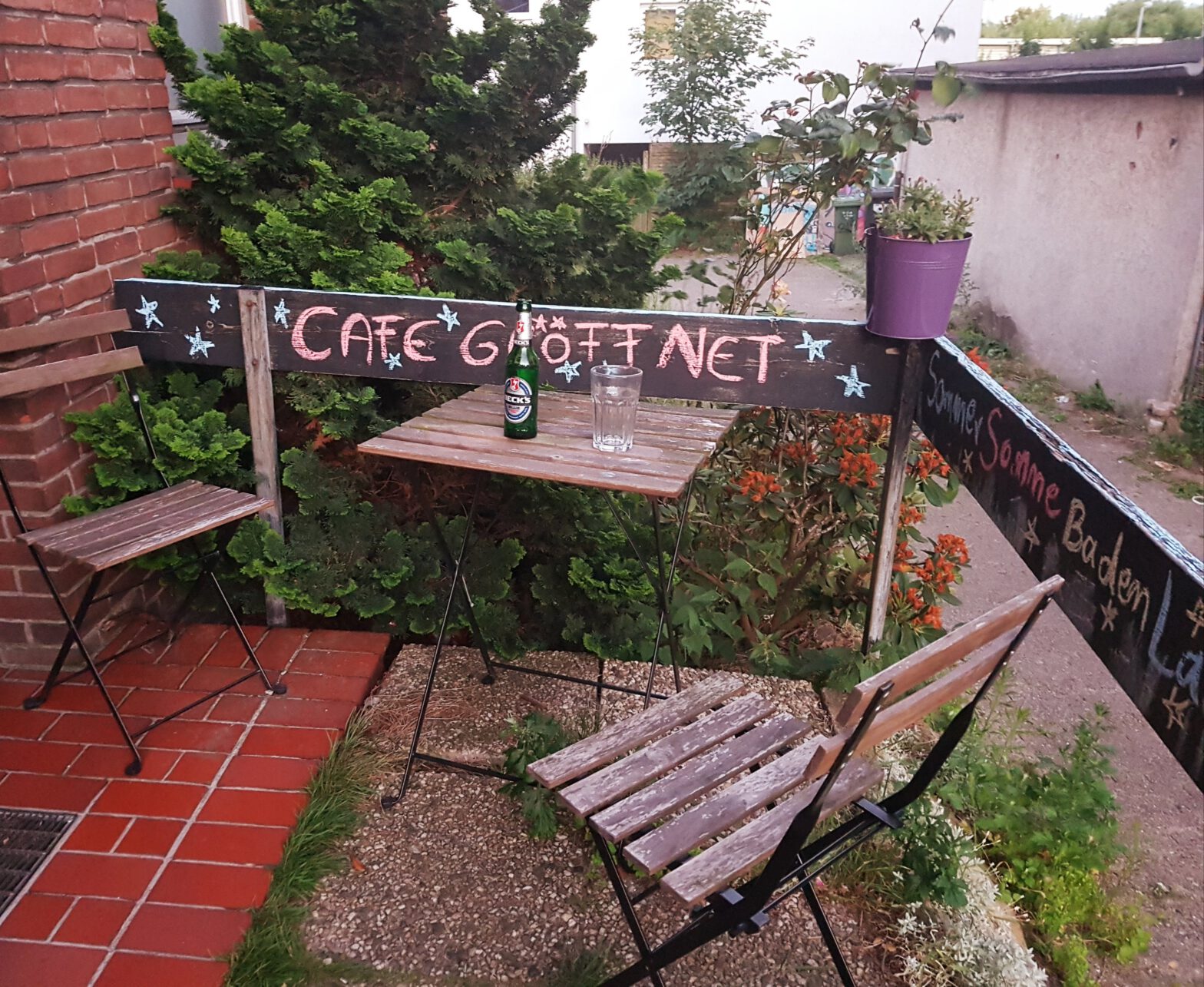 Tisch und zwei Stühle vor Haustür. An Holzbrett steht mit Kreide geschrieben: "Cafe geöffnet"