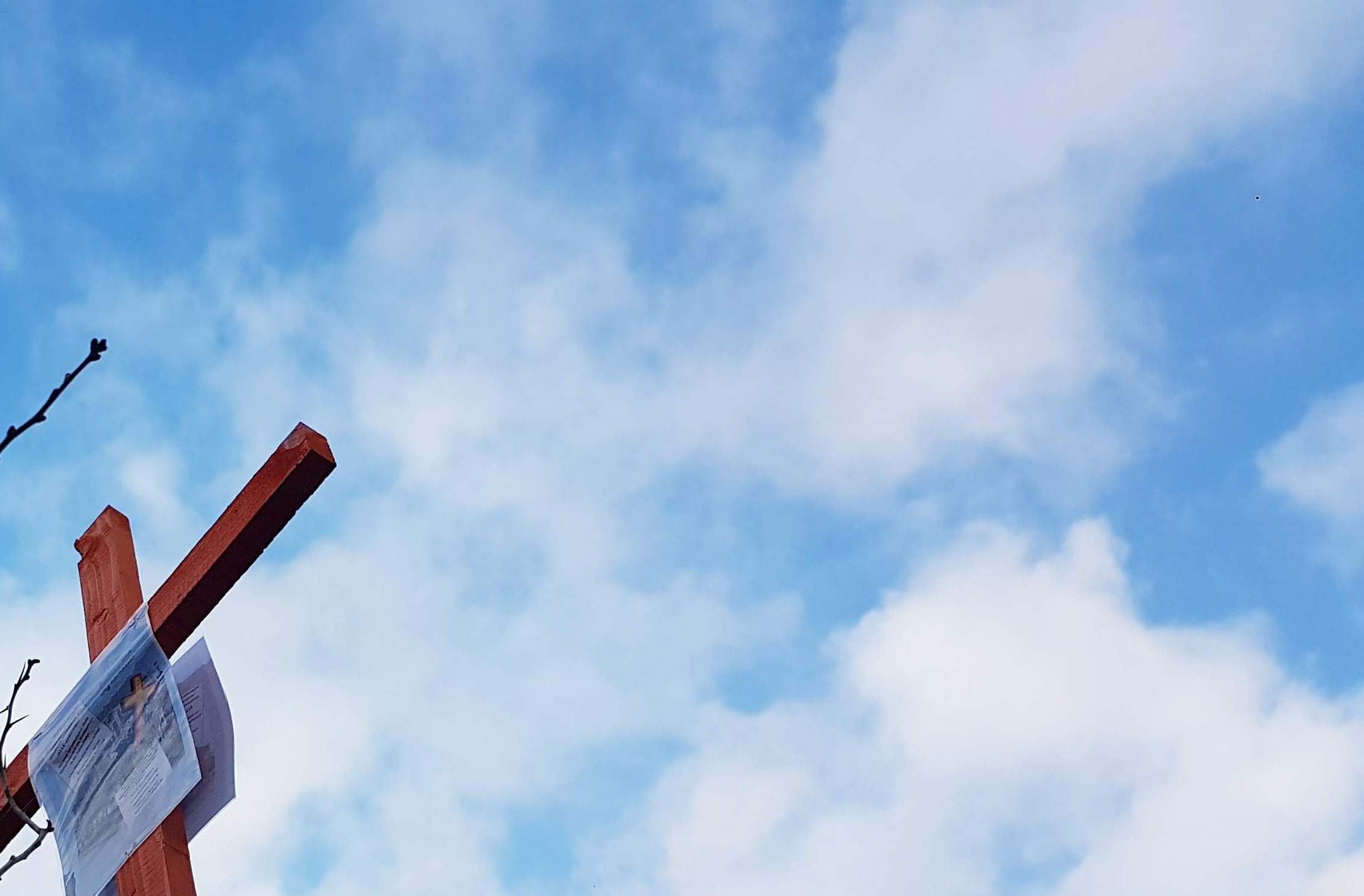 Blauer Himmel mit Wolken, in der unteren linken Ecke der obere Teil eines Holzkreuzes mit unleserlichem Zettel daran