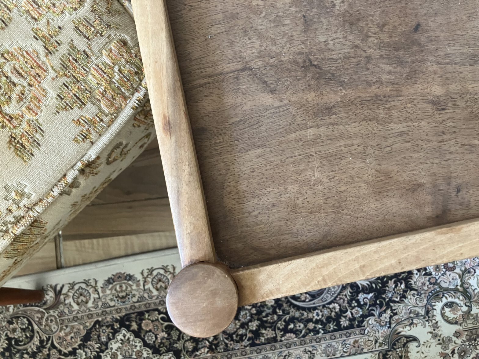 Teil einer Ecke eines Holztisches, daneben ein Polsterausschnitt eines Sessels, dunkle Töne, unten drunter ein Teil eines Teppichs mit Paisley Muster