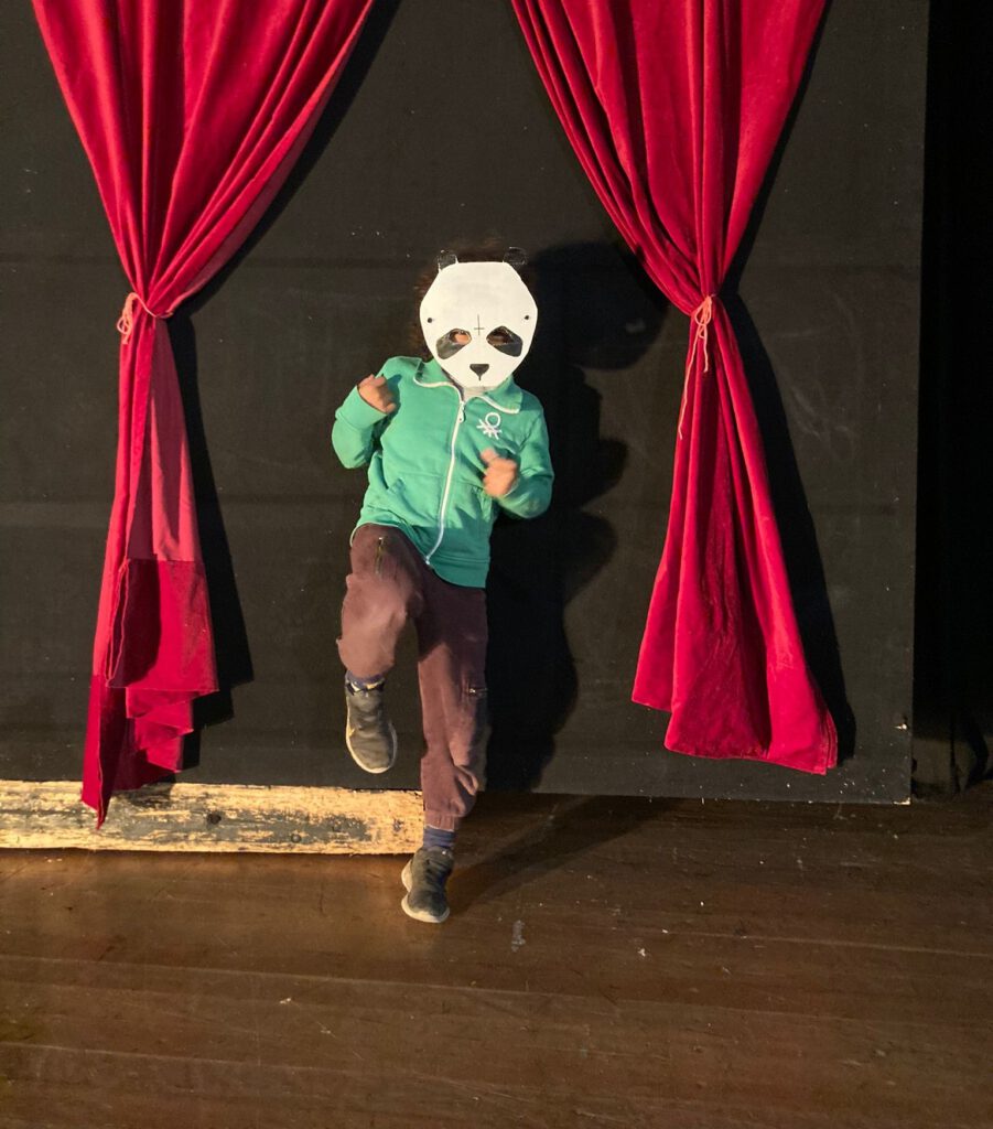 Kind mit Pandamaske aus Pappe macht Pose vor geöffnetem roten Vorhang