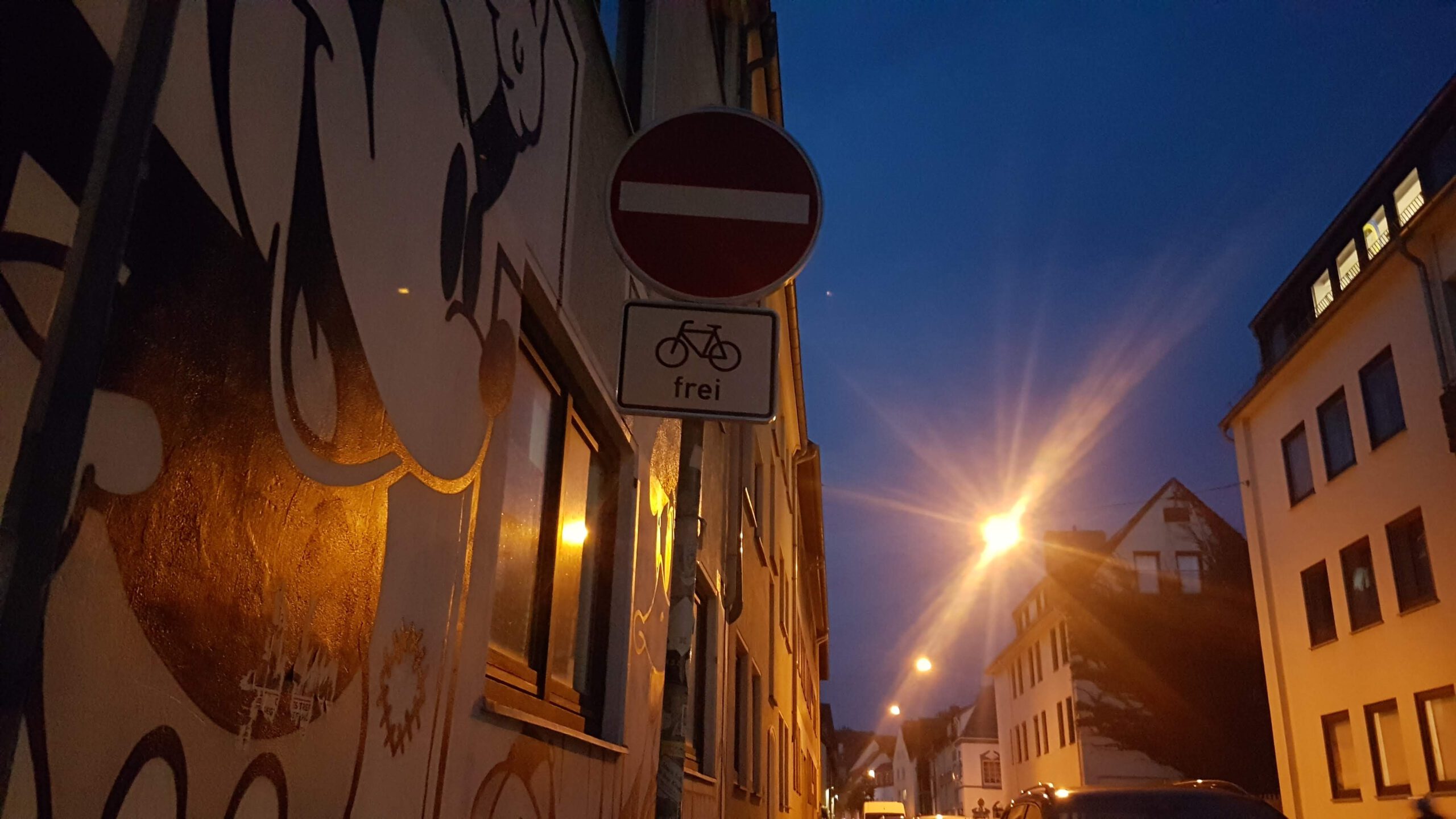 Straße mit Häuserfassaden. Direkt links auf Hauswand große gemalte Mickey Mouse. Himmel wenig zu sehen, dunkel werdend. Dunkelblau. Straßenlaternen leuchten. Licht reflektiert.