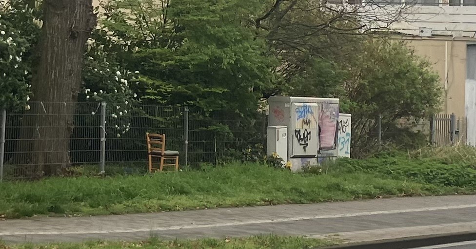 Ausschnitt von Straßenrand, links ein Baumstamm, rechts daneben ein auf Rasen abgestellter Holzstuhl, rechts daneben Stromkästen mit Graffiti